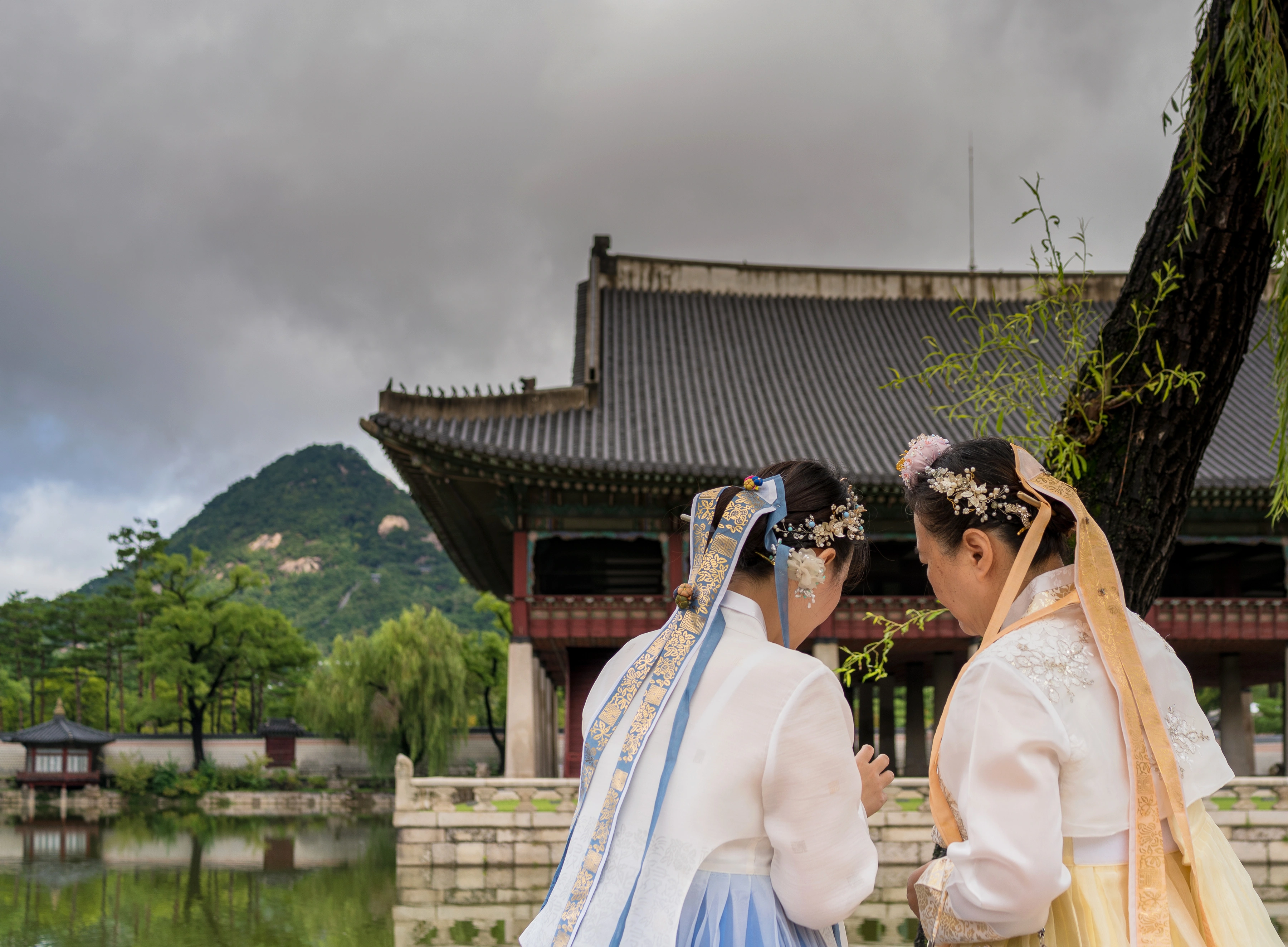 경복궁 연못과 전각을 배경으로 푸른색과 노란색 한복을 입은 두 방문객의 뒷모습